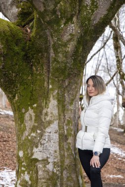 Beyaz ceketli bir kadın ormanda yosun kaplı bir ağacın yanında duruyor.