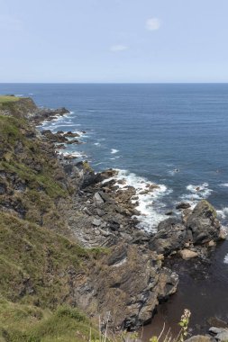 Arka planda okyanus olan kayalık kıyı şeridi, açık mavi gökyüzünün altında beyaz bulutlar.