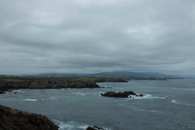 Bulutlu bir gökyüzünün altında engebeli suları olan kayalık kıyı şeridi ve arka planda tepe manzarası var.