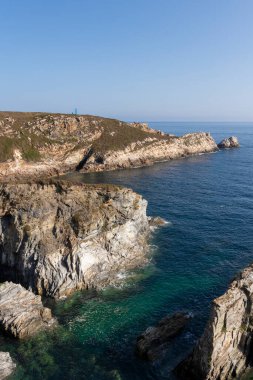 Açık mavi suları ve uzakta küçük bir deniz feneri olan kayalık bir sahil şeridi.
