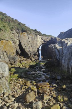 Doğal bir kemer oluşumuna sahip kayalık kıyı şeridi, açık mavi gökyüzünün altında yeşillik ve yosunlarla kaplanmış içinden akan bir dere.