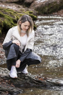 Beyaz ceketli bir kadın ormanda bir derenin kenarında çömelmiş duruyor.