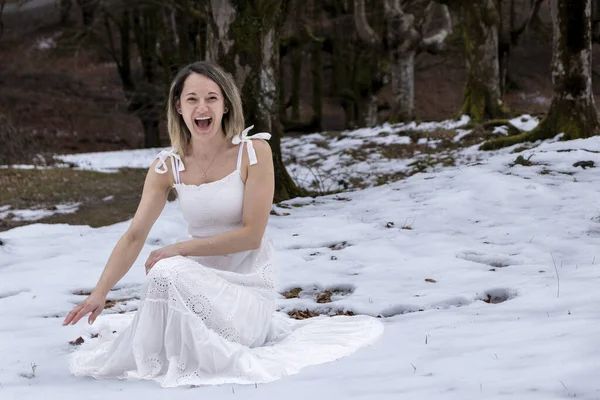 woman in a white, flowing dress with a lace pattern is sitting on a rock in a snowy forest, creating a peaceful and serene mood