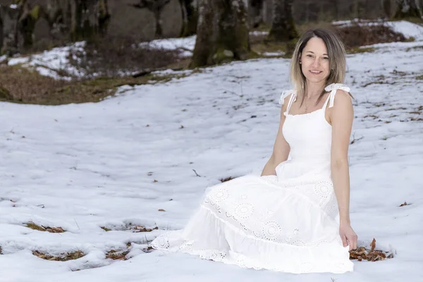 person in a long, flowing white dress sitting on a rock in a snowy forest with trees in the background