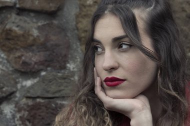 Genç bir kadın, kahverengi saçlı, kırmızı giyinmiş, dokulu taş bir arka planda.