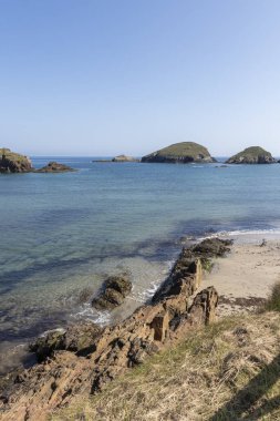 Berrak gökyüzü, kayalık kıyı, sakin sular ve parlak gökyüzünün altındaki uzak yeşil adalar ile sakin bir sahil manzarası.