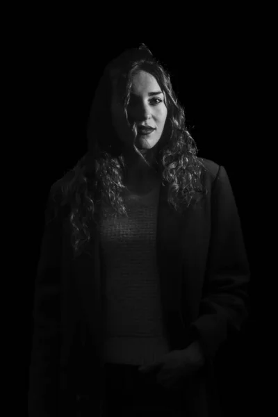 woman in a sweater and jacket is highlighted against a dark backdrop, in a moody black and white photo