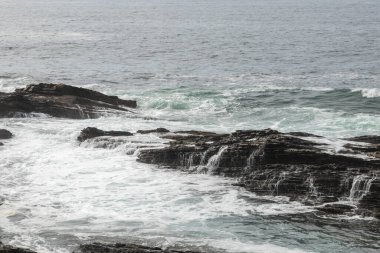 Dalgalar karanlık, pürüzlü kayalara çarpar. Denizin gücü ve güzelliği aşikardır.