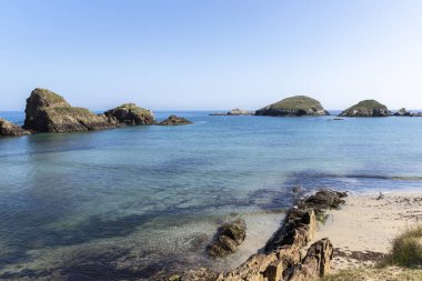 Berrak mavi suları olan sakin bir sahil, berrak bir gökyüzünün altında kayalık oluşumlar, sükunet ve sükunet hissi uyandırıyor.