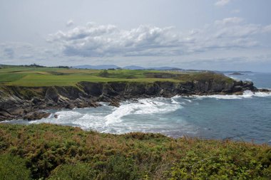 Yeşil tarlaları, kayalık uçurumları ve parçalı bulutlu bir gökyüzünün altında canlı mavi bir okyanusu olan manzaralı kıyı manzarası.