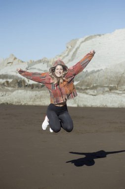 Kadın sevinçle sahilde zıplıyor, gölgesini düşürüyor, arka planda kayalık kayalıklar var.