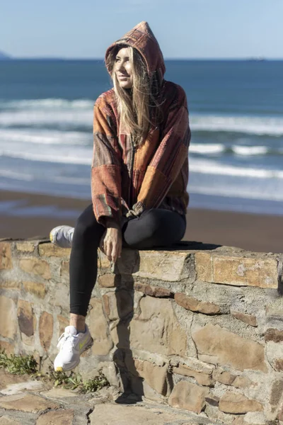 woman in a hoodie sits on a stone wall, overlooking a beach with waves rolling in under a clear blue sky