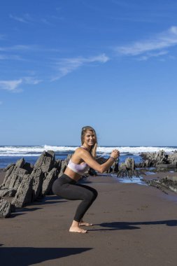 Kadın kumlu bir plajda yoga yapıyor. Arkasında kayalar ve dalgalar açık bir gökyüzünün altında.