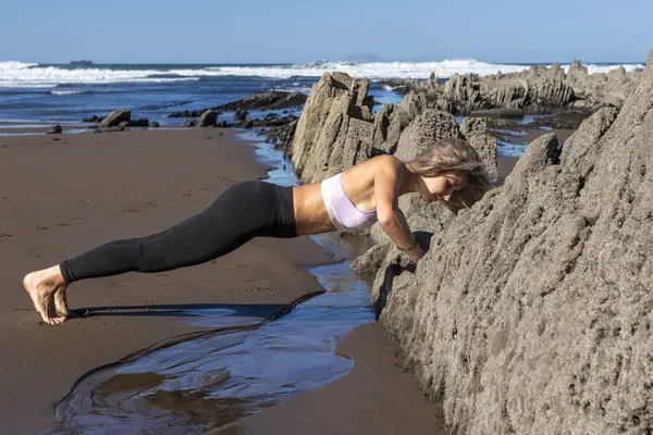 Kadın açık gökyüzünün altında kayalık oluşumlarla bir plajda şınav çekiyor.