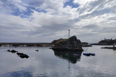Teknelerle dolu sakin bir liman, bulutlu bir gökyüzünün altında kayalık oluşumlarla çevrili, sakin ve huzurlu bir atmosfer yayıyor.