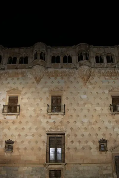 historic building with textured walls and arched structures, illuminated against the night sky. It features several windows with wrought iron grills or balconies. The architecture appears ancient and intricate
