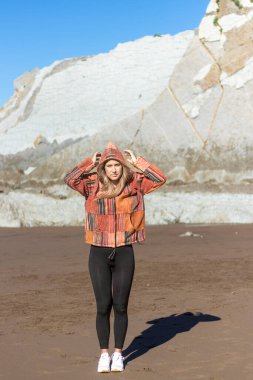 Kapüşonlu ceket giyen bir kadın kumsalda duruyor. Ceket renkli ve yamalı bir tasarımı var.