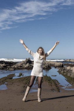Kadın plajda duruyor, beyaz bir elbise ve çizme giyiyor. Gülümsüyor ve mutlu.