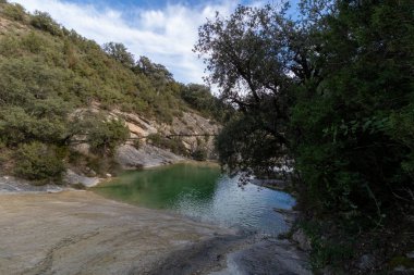 Berrak bir gökyüzünün altında yemyeşil ağaçlarla çevrili kayalık tepelerin arasına kurulmuş dingin yeşil gölet. Doğal bir güzellik yayar.