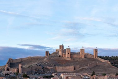 Birden fazla kulesi olan antik taş kale bulutlu bir gökyüzünün zeminine karşı kurulmuş küçük bir kasabaya bakıyor.