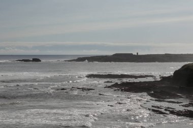 Parçalı bulutlu bir gökyüzü altında dalgaların çarptığı kayalık kıyı şeridi doğanın dingin ama ehlileştirilmemiş güzelliğini simgeliyor.
