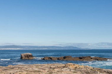 Kayalık bir kıyısı, sakin bir denizi ve berrak bir gökyüzünün altındaki uzak dağları olan huzurlu kıyı manzarası.