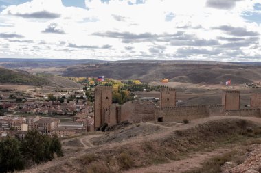 Üç bayraklı antik taş kale, bir vadide bir kasabaya tepede yuvarlanan tepeler ve bulutlu bir gökyüzü