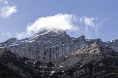 Mavi gökyüzünün altında beyaz bulutlarla vurgulanmış görkemli bir dağ zirvesi.