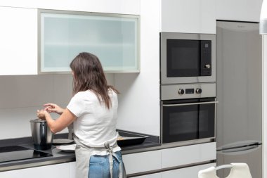 Modern bir mutfakta fırında tencereyle yemek pişiren beyaz bluz ve kot pantolon giyen biri.