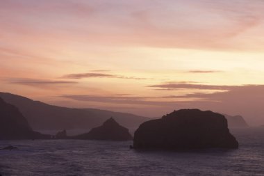 Okyanusun üzerinde sakin bir gün batımı, siluetli kayalık adalar ve sıcak pembe ve turuncu renkler gökyüzünde