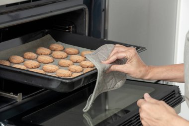 Fırından bir tepsi kurabiye çıkarmak için fırın eldiveni kullanan birinin elleri.