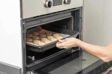 Kurabiyeleri fırından çıkaran kişi. Fırın gümüş ve siyah. Kurabiyeler fırın tepsisinin üstünde.