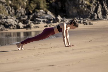 Kadın plajda şınav çekiyor. Kumsal kayalık ve su arka planda.