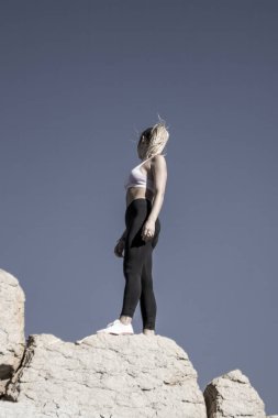 Atletik bir kadın kayalık bir zirvede kendine güvenle duruyor, açık mavi gökyüzüne bakarak.