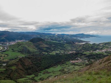 Yeşil tepelerin, kıyı köylerinin ve denizin yakınındaki bulutlu bir gökyüzünün altındaki uzak dağların panoramik manzarası.