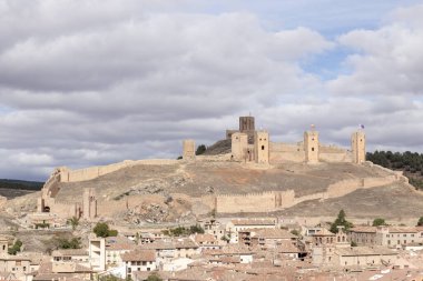 Bulutlu bir gökyüzünün altındaki tarihi bir köye bakan güçlendirilmiş surları olan görkemli bir ortaçağ kalesi.