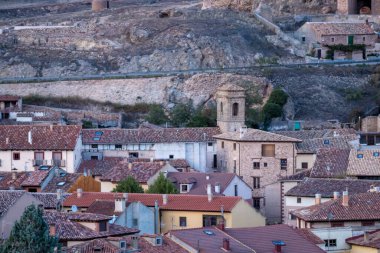 Taş kilise kulesi ve kayalık bir tepenin üzerinde kırmızı kiremitli çatıları olan tarihi bir köyün yakın görüntüsü.