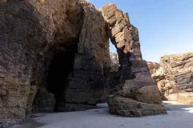 Parlak mavi gökyüzünün altında kumlu bir sahilde doğal kaya kemeri ve engebeli uçurumlar..