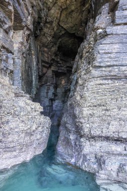 Narrow rocky crevice with crystal-clear turquoise water leading into a cave-like formation. clipart