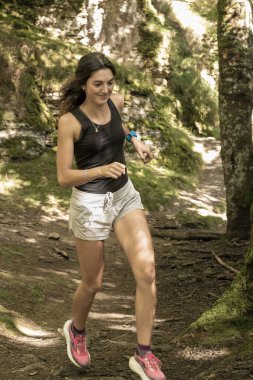 Güneşli bir günde, yosunlu kayalar ve ağaçlarla çevrili ormanda koşan gülümseyen bir kadın..