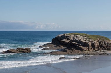 Coastal rock formation surrounded by ocean waves and a clear blue sky on a sunny day. clipart