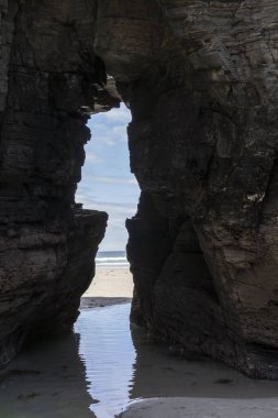 Kumlu bir sahile açılan doğal kaya geçidi. Okyanus dalgaları görülebilir. Çerçevesi koyu kayalıklarla çevrili.