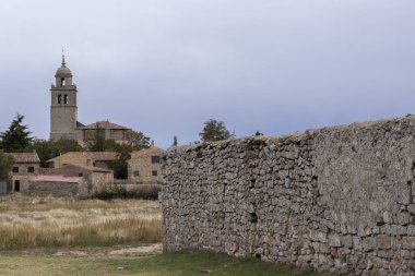 Rustic village with stone houses and a prominent church tower, surrounded by fields and historic stone walls under an overcast sky. clipart