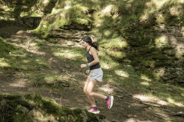 Ağaçların arasından süzülen yemyeşil ve güneş ışığıyla çevrili bir ormanda koşan bir kadın açık havada aktif bir yaşam tarzının tadını çıkarıyor.