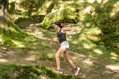 Genç bir kadın ormanda yokuş aşağı koşuyor, atletik giysiler ve spor ayakkabılar giyiyor. Güneş ışığı ağaçların arasından süzülüp yeşil yosunlu araziyi aydınlatıyor.