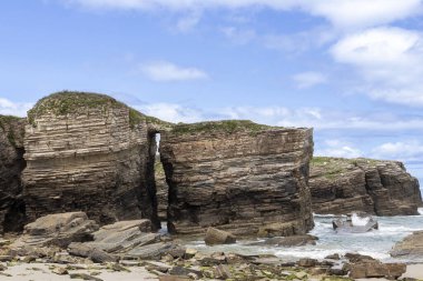 Katmanlı tortulu kayalık oluşumları ve okyanusa karşı doğal bir kemer duruşu. Dalgalar mavi gökyüzünün altındaki engebeli kıyı şeridine çarpar.