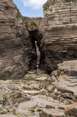 Eroded rock formations with a natural archway along a rugged coastline. Layers of sedimentary rock, scattered stones, and green vegetation create a dramatic landscape clipart