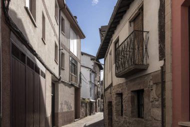 Binanın kenarında balkon olan dar bir ara sokak. Balkon boş. Sokak binalarla dolu ve taş bir duvarı var.