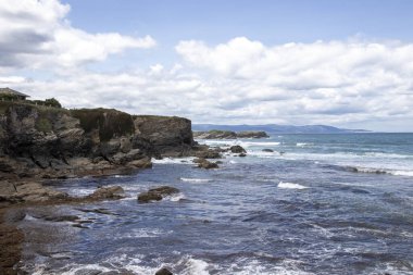 Kıyı kayalıkları berrak mavi denizle buluşuyor, dalgalar yavaşça kırılıyor ve dağlar sisli bir mesafede görünüyor.