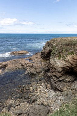 Küçük mağarası, gelgit havuzları ve güneşli bir gökyüzünün altında berrak mavi okyanusa bakan dağınık çakıl taşları olan kayalık bir sahil kayalığı.